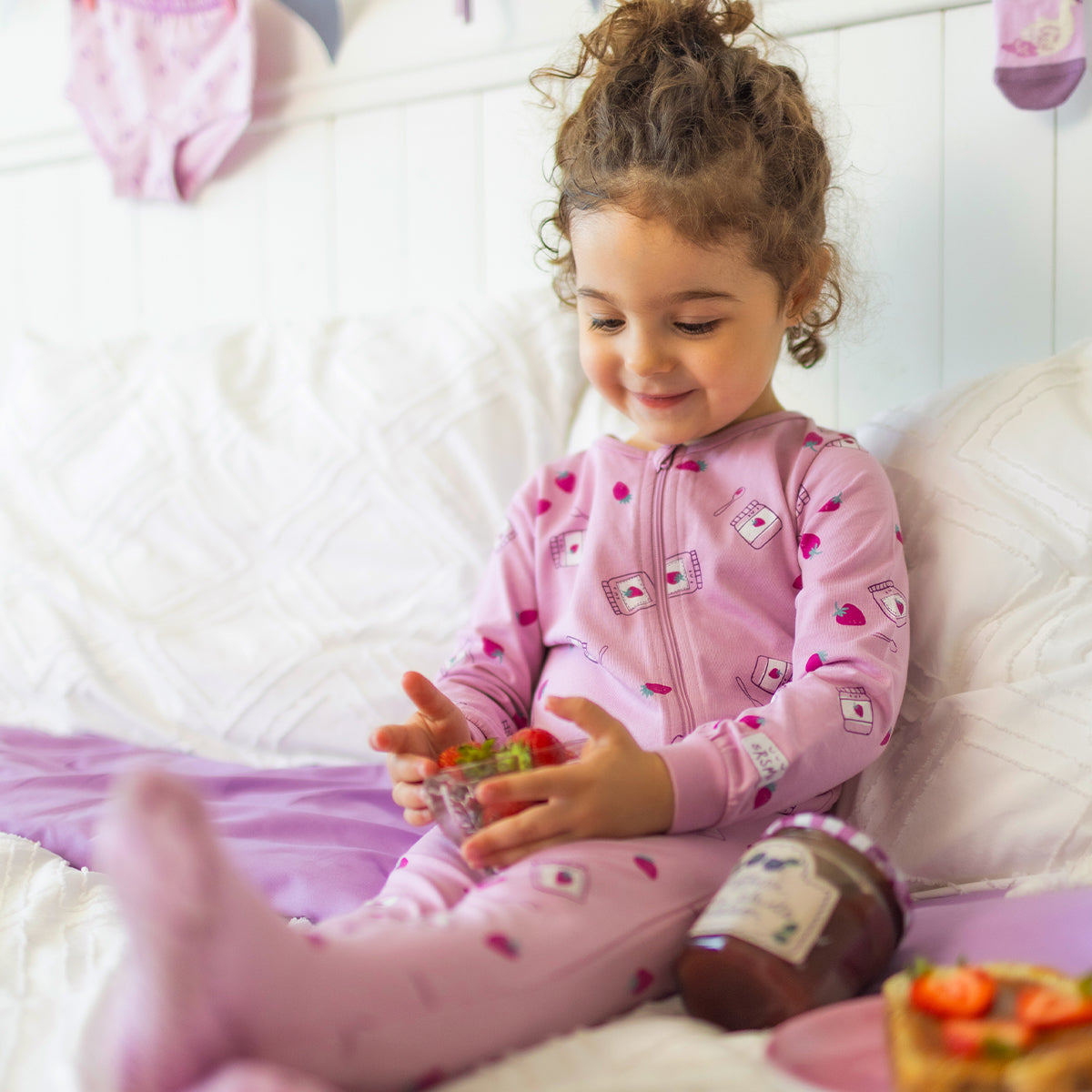 Pyjama une pièce mauve à petits motifs pour bébé en coton doux, porté par un bébé assis sur un lit, avec fermeture éclair pleine longueur et imprimé de fraises et pots de confiture.
