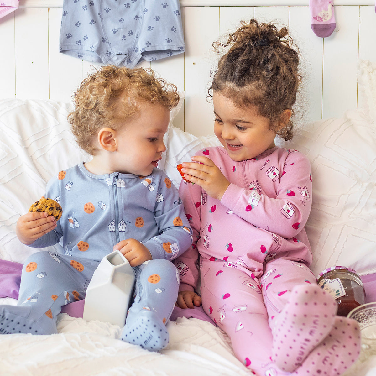 Pyjama une pièce mauve à petits motifs pour bébé et pyjama une pièce bleu à petits motifs pour bébé en coton doux, portés par deux bébés assis sur un lit