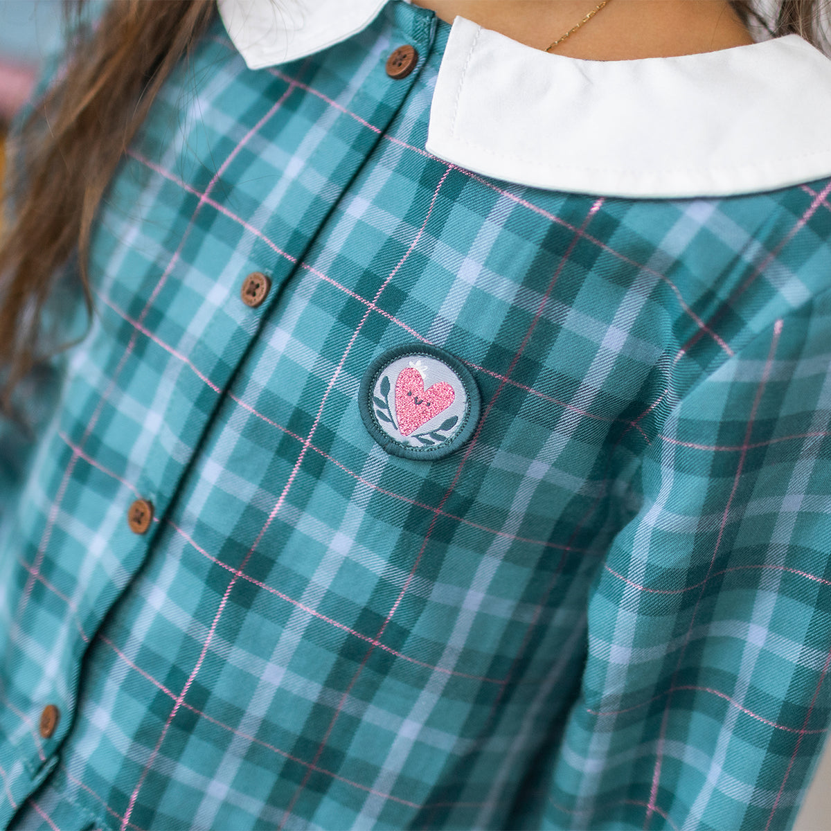 Gros plan d'une robe turquoise à carreaux en flanelle pour enfant, avec col blanc, boutons effet bois et écusson cœur brodé sur la poitrine.