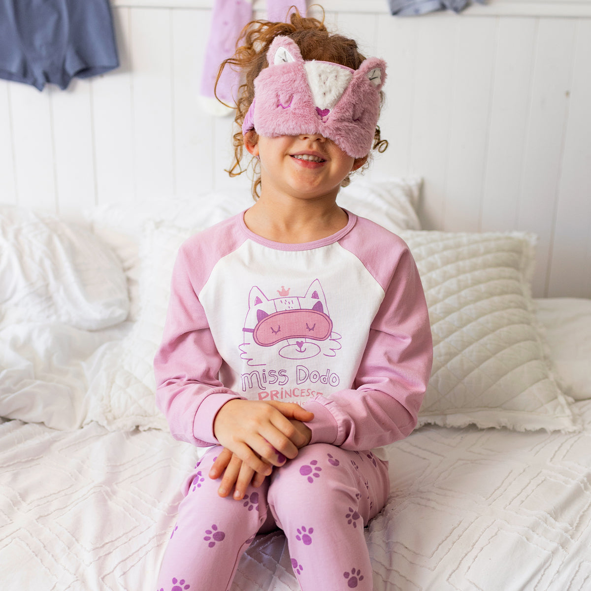 Enfant portant le pyjama deux pièces rose à illustration de chat en coton doux, assis sur un lit avec un masque de sommeil en forme de chat assorti