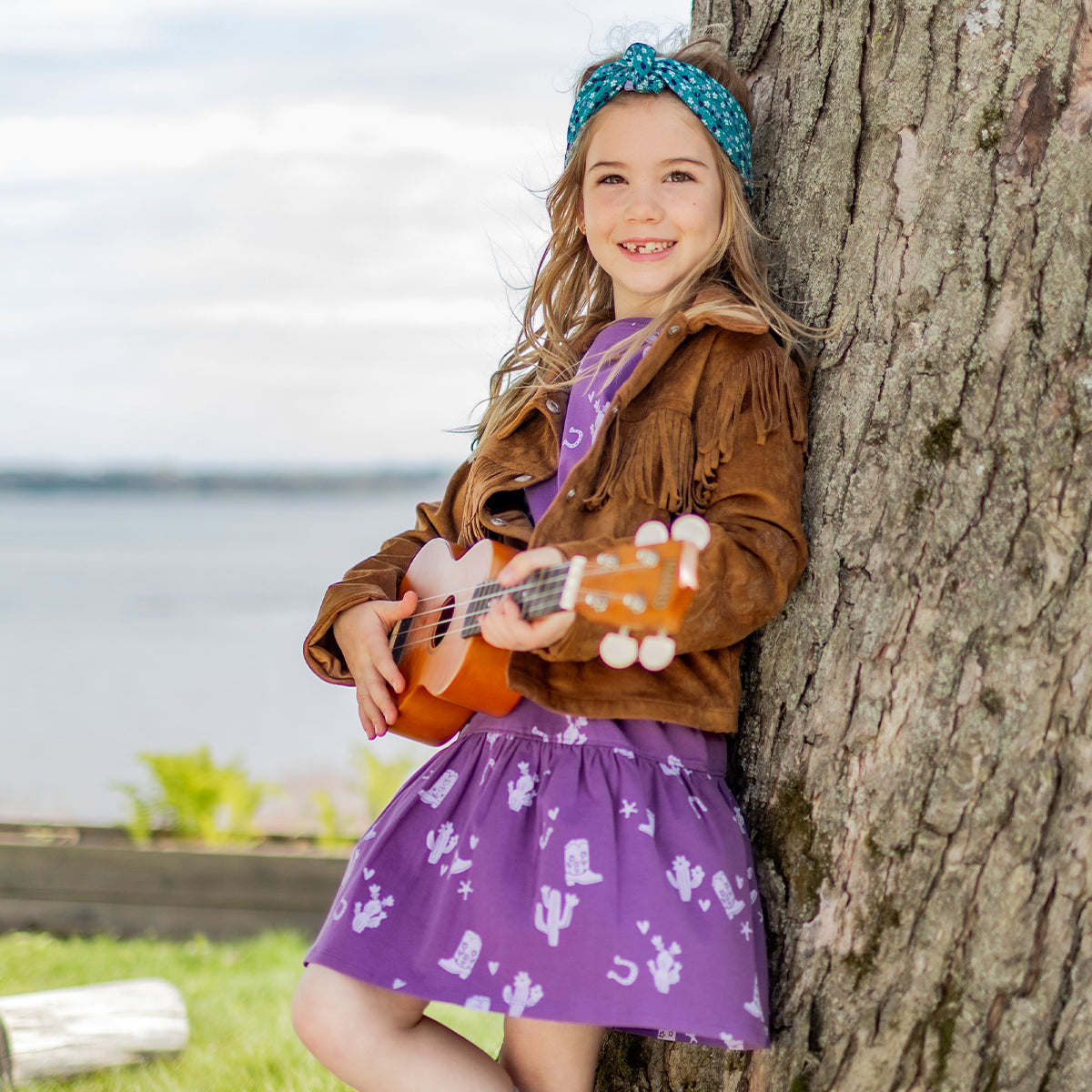 Fille souriante portant la robe mauve à motifs western et une veste à franges brune, jouant du ukulélé près d’un arbre, en plein air.