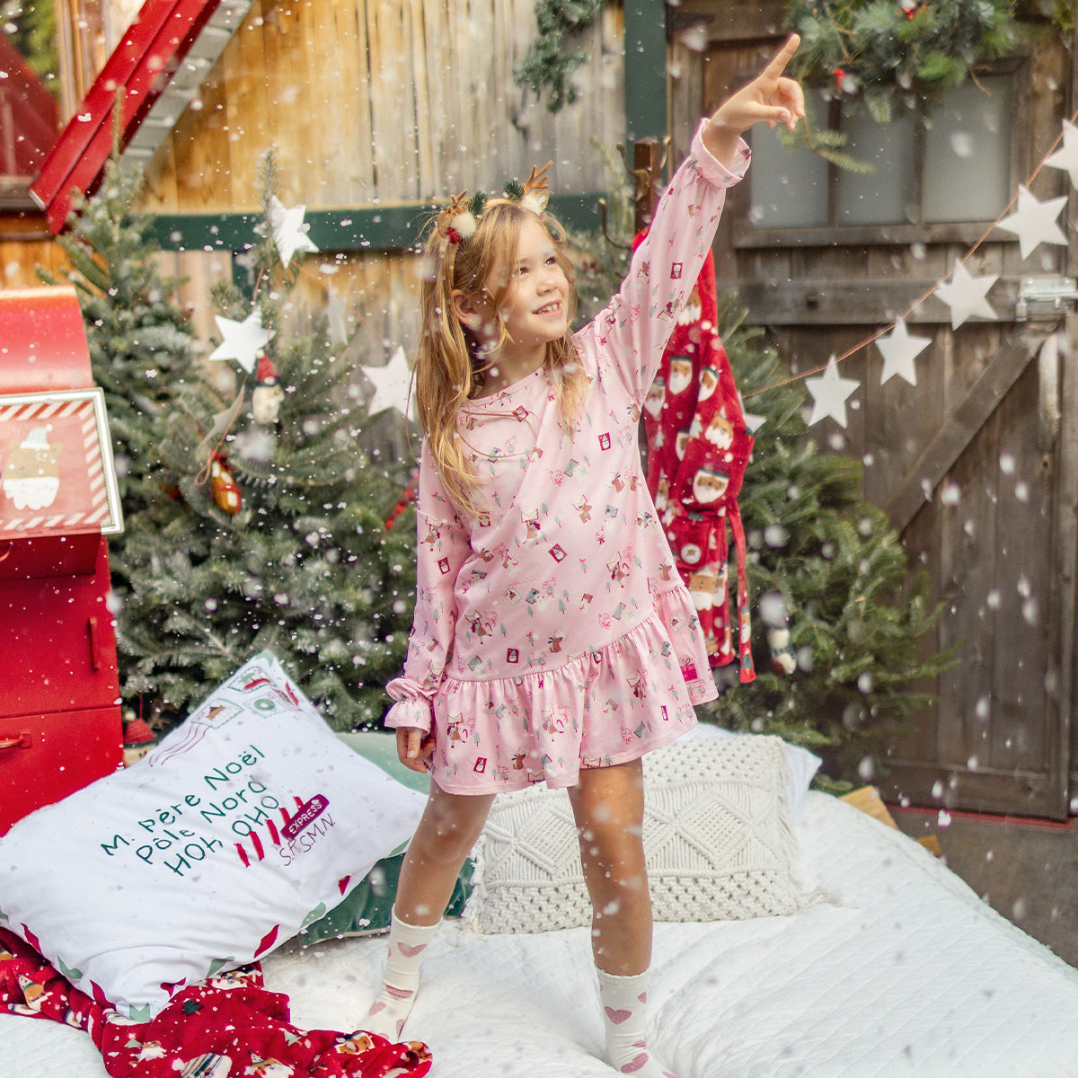 Fillette en nuisette rose à motifs des Fêtes jouant sous la neige, entourée de décorations de Noël et d’un grand sapin illuminé. - Souris Mini