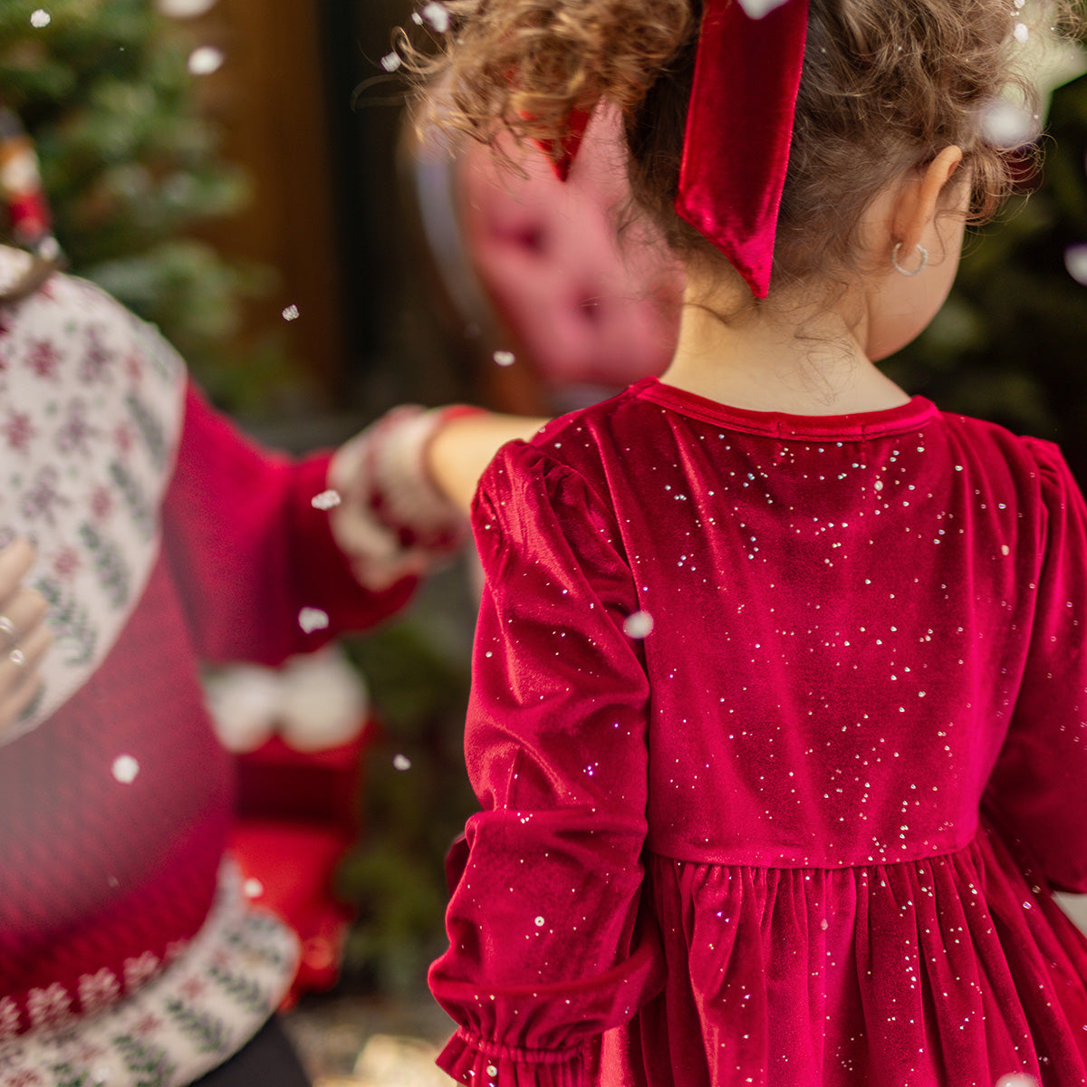 Zoom de dos d'une fillette portant une robe rouge en velours scintillant avec un nœud, debout dans un décor de Noël chaleureux, collection Souris Mini.