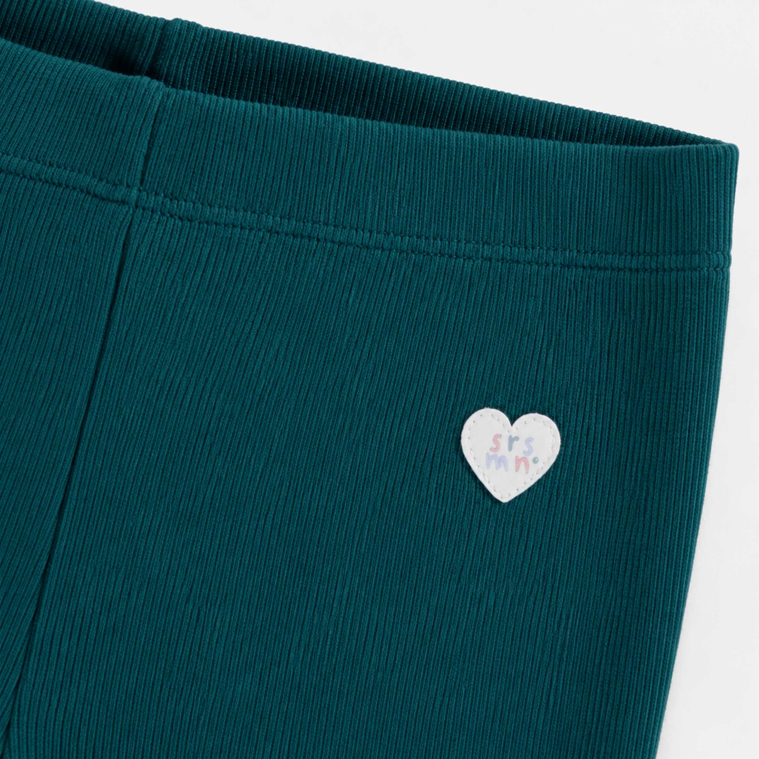 Gros plan sur un legging côtelé vert pétrole pour bébé fille, mettant en valeur la taille élastique et un petit écusson cœur cousu à la hanche.