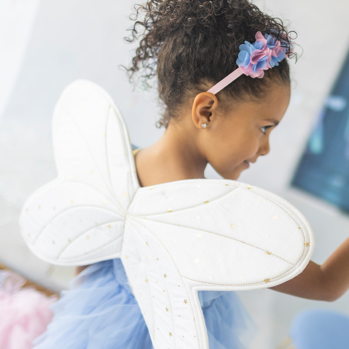 Petite fille portant des ailes d’ange blanches avec motifs étoilés dorés et un serre-tête fleuri rose et bleu, déguisement enfant féerique.