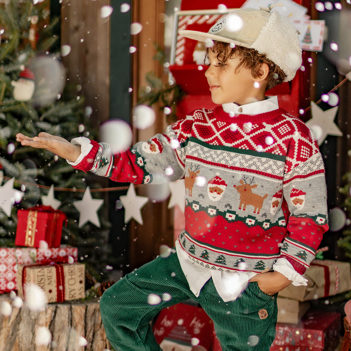 Petit garçon portant un chandail rouge de Noël en tricot avec des motifs de rennes et de Père Noël, posant dans un décor festif enneigé.