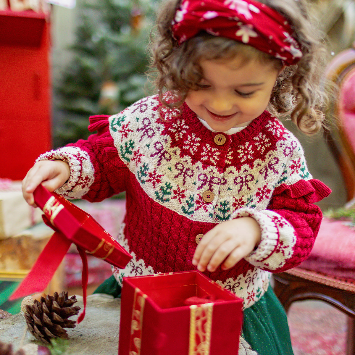 Fillette souriante ouvrant un cadeau, vêtue d’un chandail rouge et blanc en tricot festif avec motifs de Noël, collection Souris Mini.