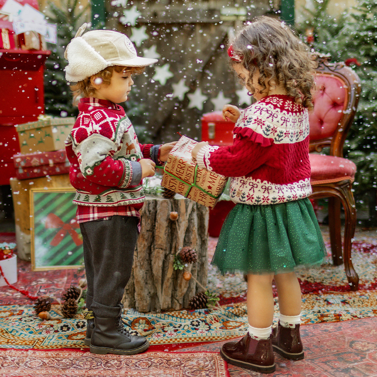 Deux enfants vêtus de chandails en tricot festifs s’échangent un cadeau dans un décor de Noël magique, collection Souris Mini.