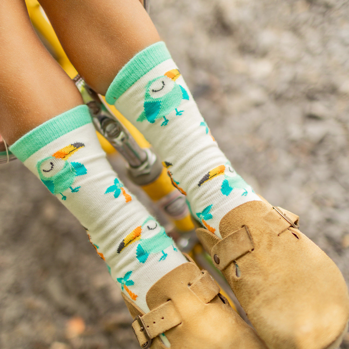 Gros plan sur des chaussettes crème à motifs de toucans turquoise et orange portées par un enfant assis sur un vélo jaune, avec des chaussures beige en suède. - Souris Mini