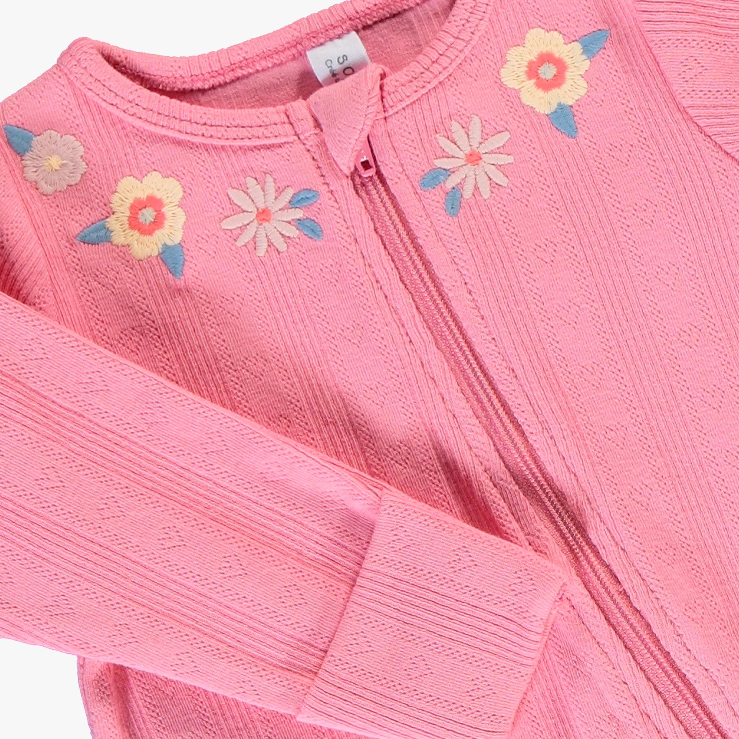 Gros plan sur les broderies florales et la fermeture zippée d’un pyjama bébé fille rose.