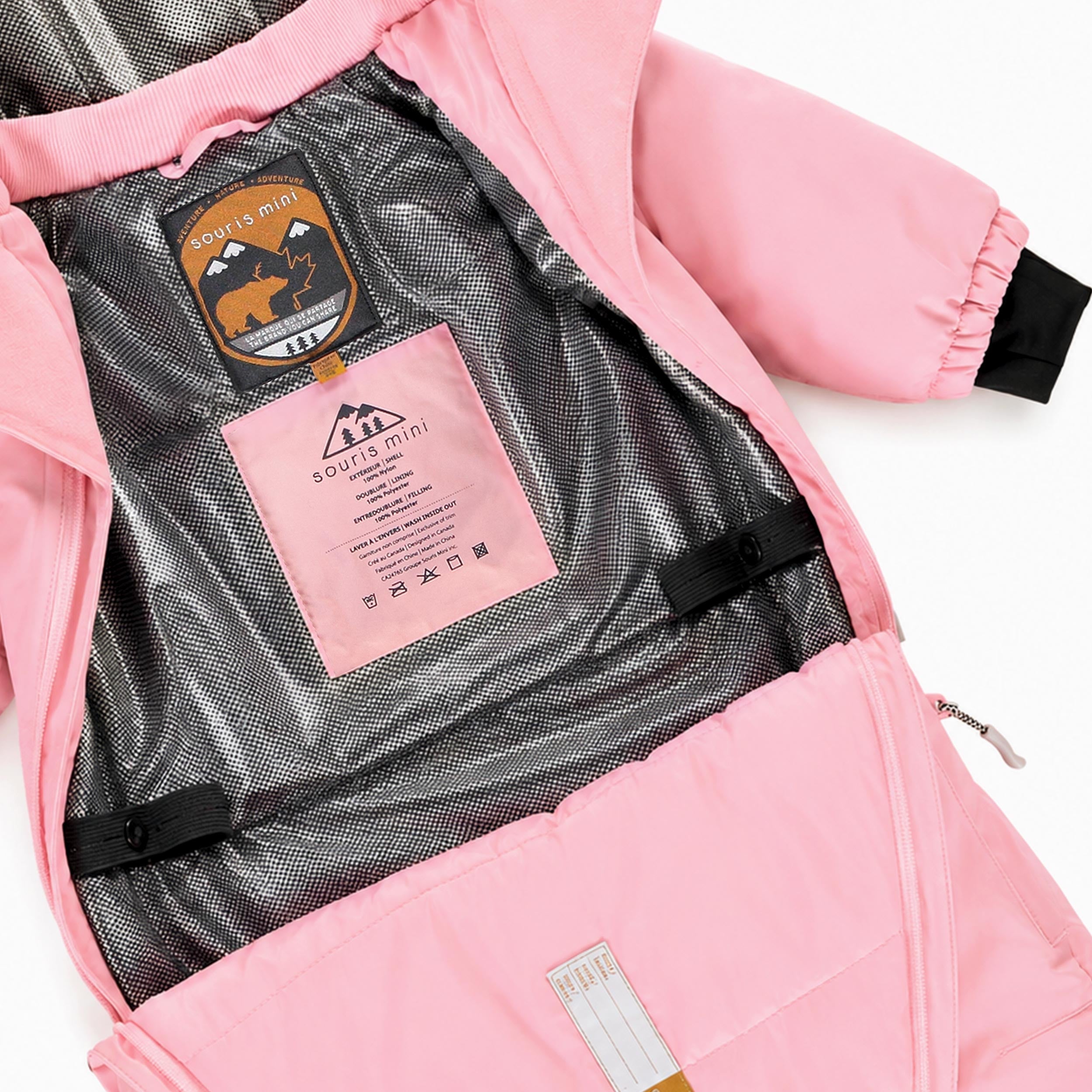 Détail intérieur d’un habit de neige pour bébé rose pâle montrant la doublure thermique réfléchissante, les bretelles de maintien et les étiquettes d’identification.