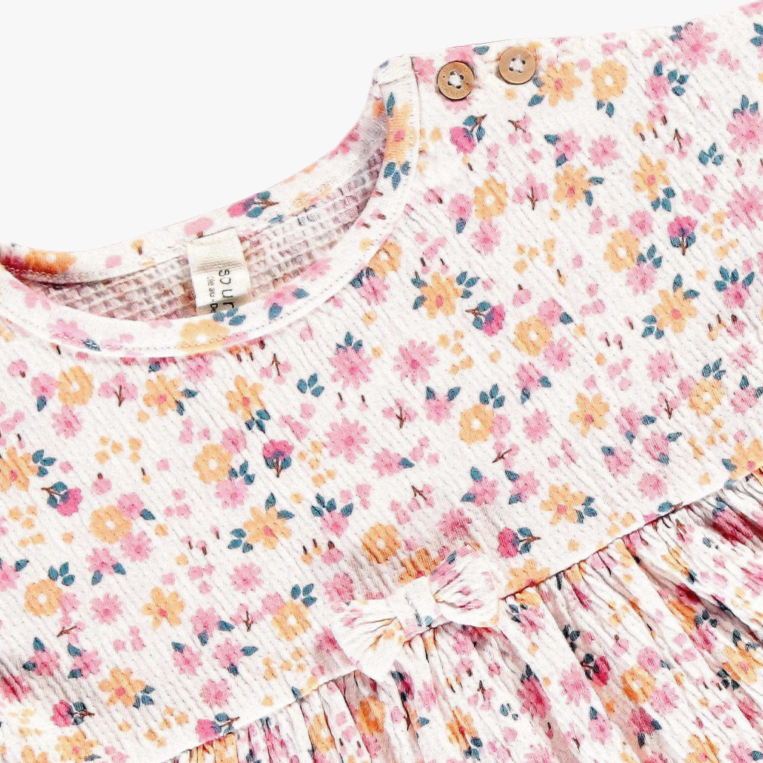 Détail d’une robe bébé fille à imprimé floral avec boutons décoratifs à l’épaule et nœud à la taille.