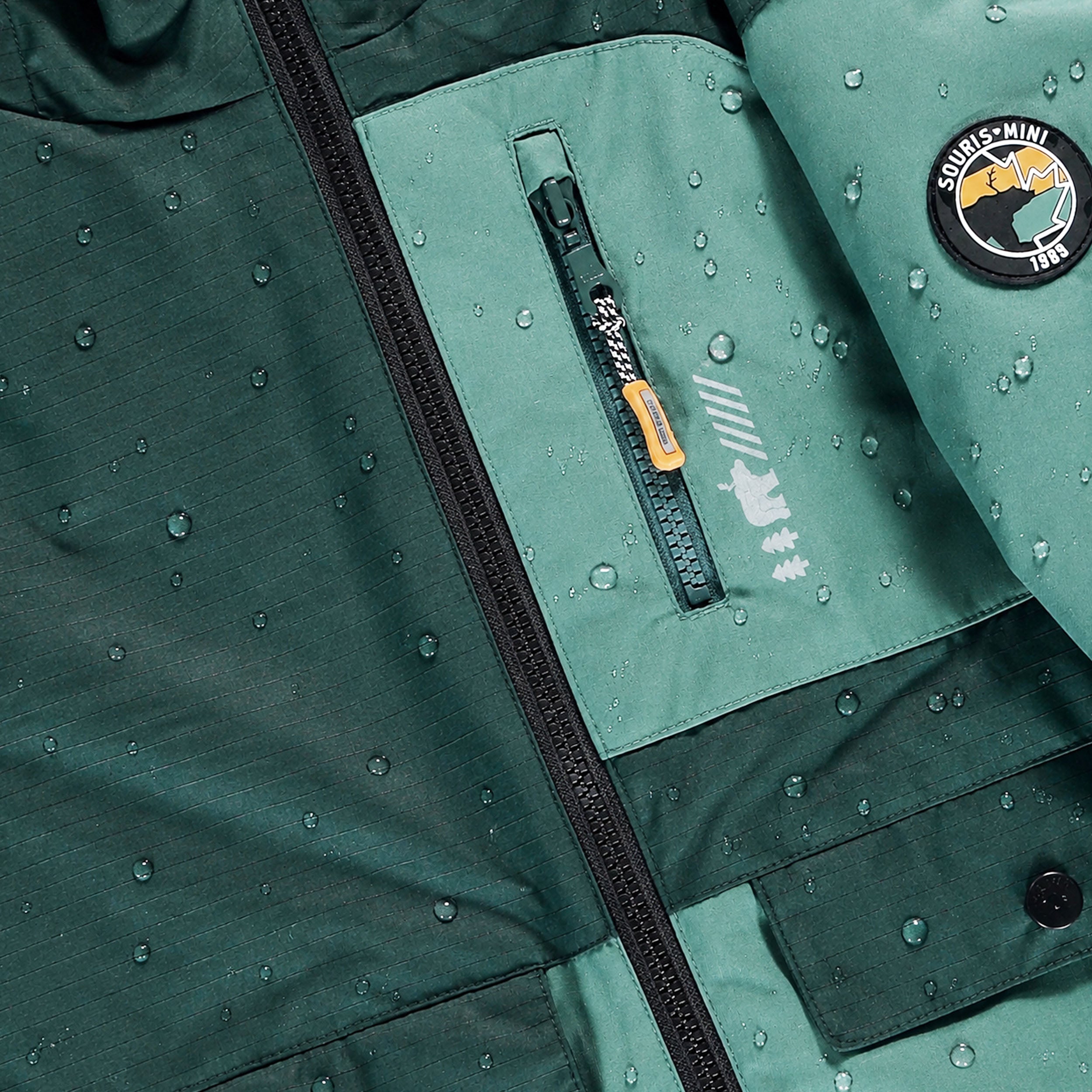 Détail d’une veste de ski enfant imperméable verte avec gouttes d’eau et poche zippée