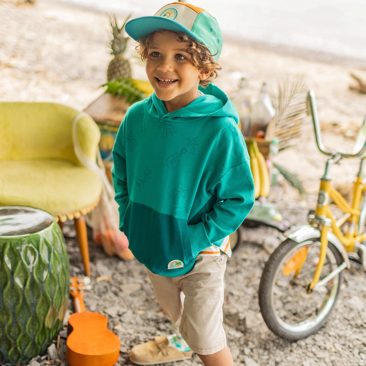 Enfant souriant portant un chandail à capuchon turquoise avec motifs de ukulélés et palmiers, une casquette bicolore et un short beige, dans un décor tropical avec vélo jaune. - Souris Mini