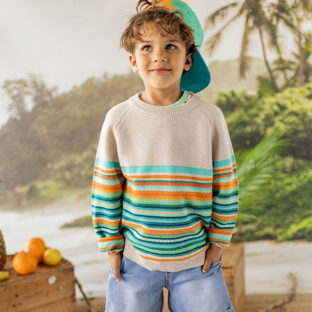 Enfant portant un chandail rayé turquoise, orange et beige, accompagné d’une casquette turquoise portée à l’envers, devant un décor tropical avec palmiers et fruits. - Souris Mini