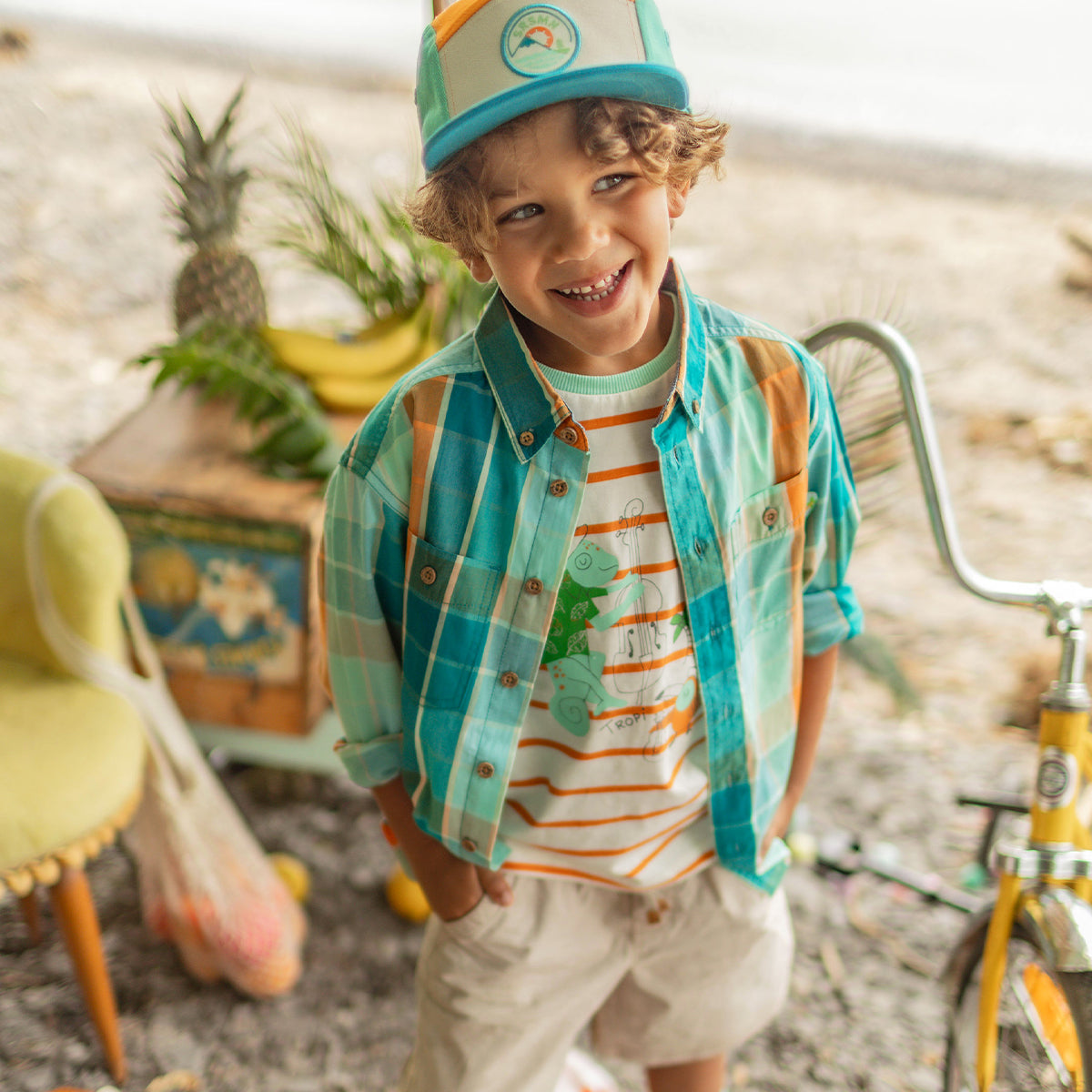 Enfant souriant portant une chemise à carreaux turquoise et orange ouverte sur un t-shirt rayé à illustration tropicale, avec une casquette bicolore, debout près d’un vélo jaune dans un décor de plage. - Souris Mini