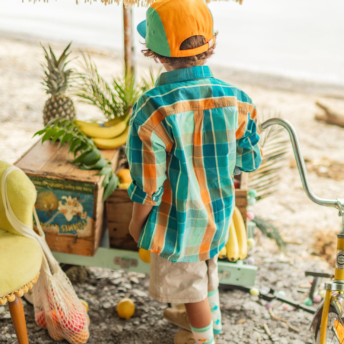 Enfant vu de dos portant une chemise à carreaux turquoise et orange et une casquette bicolore, debout près d’un présentoir de fruits tropicaux au bord de la plage. - Souris Mini