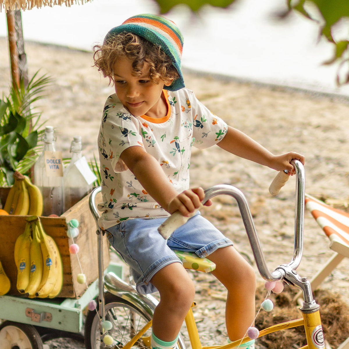 Photo estivale d’un enfant sur un vélo jaune, vêtu d’un chapeau en crochet rayé et d’un t-shirt tropical, devant une charrette de fruits au bord de la plage. - Souris Mini