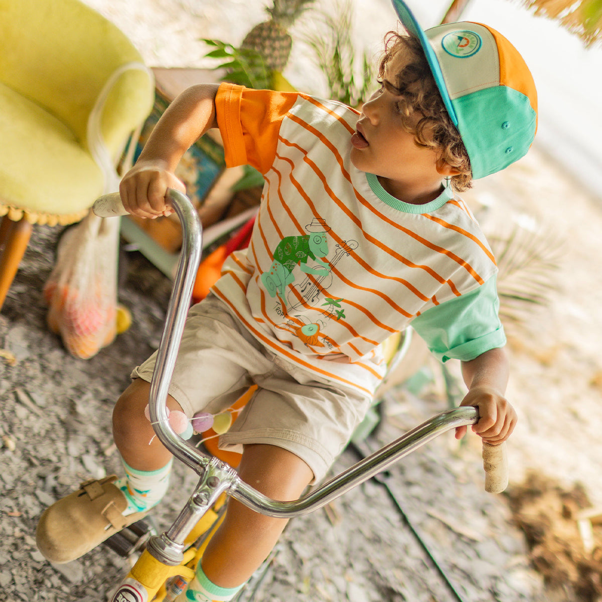 Enfant sur un vélo jaune, portant un t-shirt crème à rayures orange avec illustration tropicale, une casquette turquoise et orange, un short beige et des chaussettes colorées. - Souris Mini