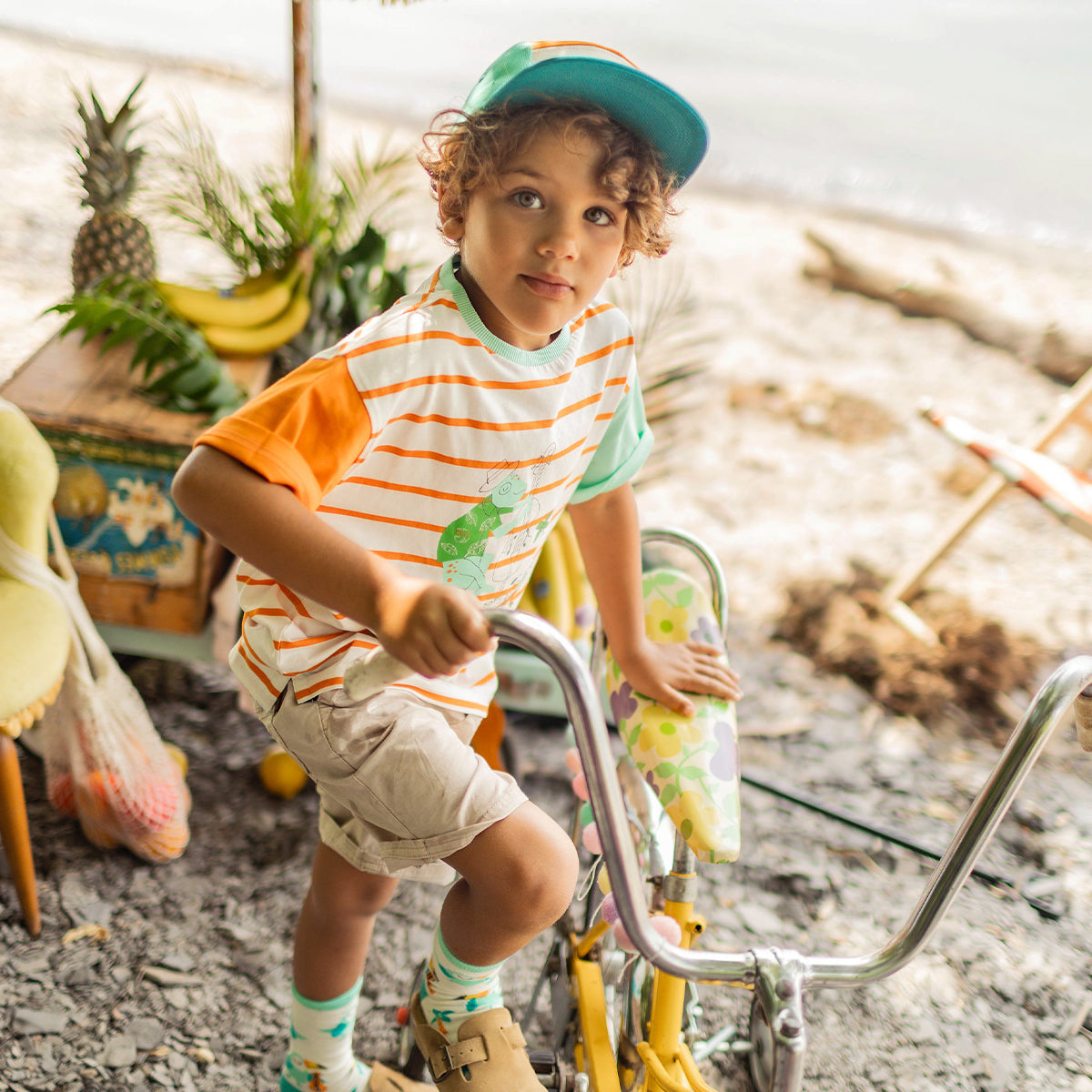 Enfant assis sur un vélo jaune à la plage, portant un t-shirt crème rayé orange avec illustration tropicale, une casquette turquoise et un short beige. - Souris Mini