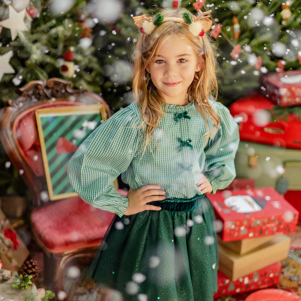 Petite fille vêtue d’une blouse verte à carreaux et d’une jupe scintillante posant dans un décor festif de Noël avec sapins et cadeaux.