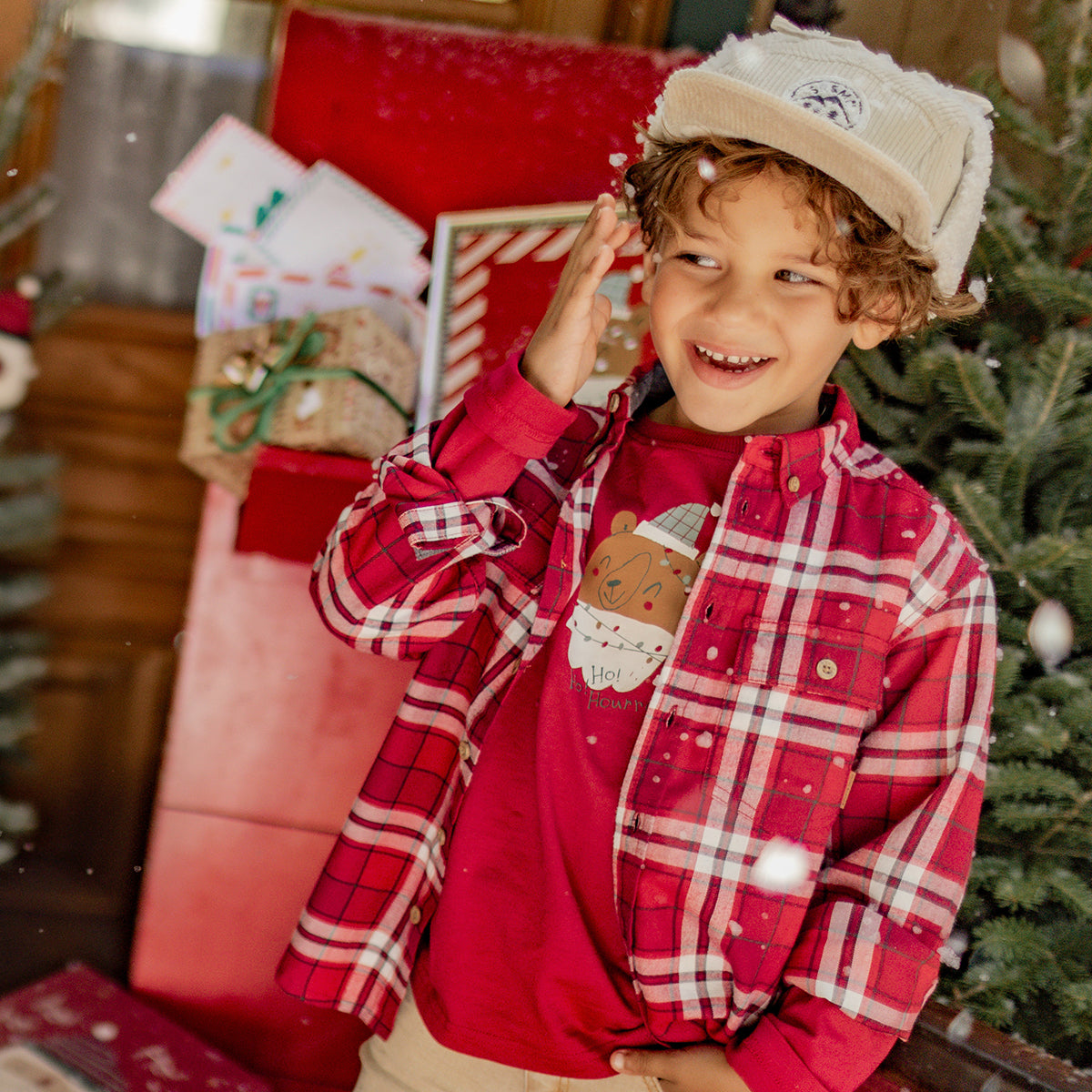 Petit garçon souriant portant une chemise rouge à carreaux et un t-shirt illustré d’un motif des Fêtes, dans un décor de Noël chaleureux.