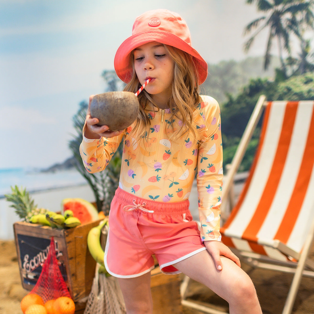 Fillette portant un rashguard orange à motifs fruités et un short rose, buvant dans une noix de coco sur un décor tropical.