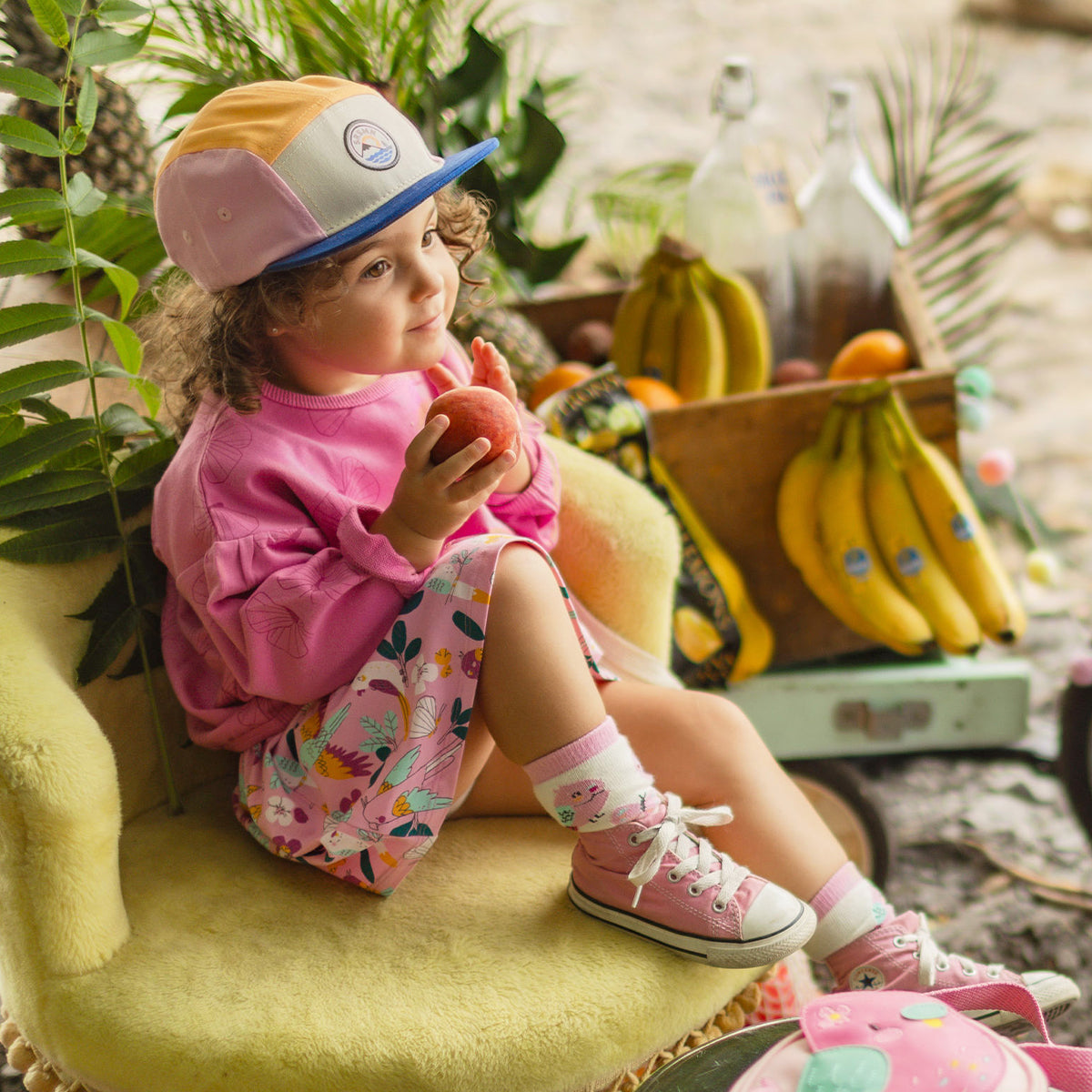 Fillette assise sur une chaise jaune portant un chandail rose et une jupe à motifs tropicaux, tenant une pomme dans un décor estival rempli de fruits.