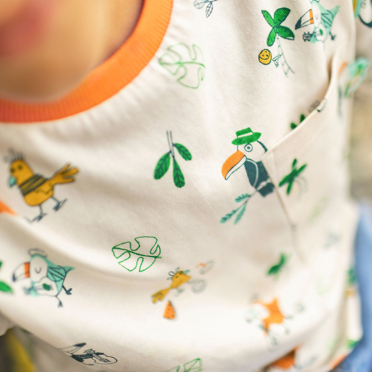 Gros plan sur un t-shirt crème à motifs tropicaux avec petits oiseaux et feuillages verts, bordure de col orange et petite poche sur la poitrine.