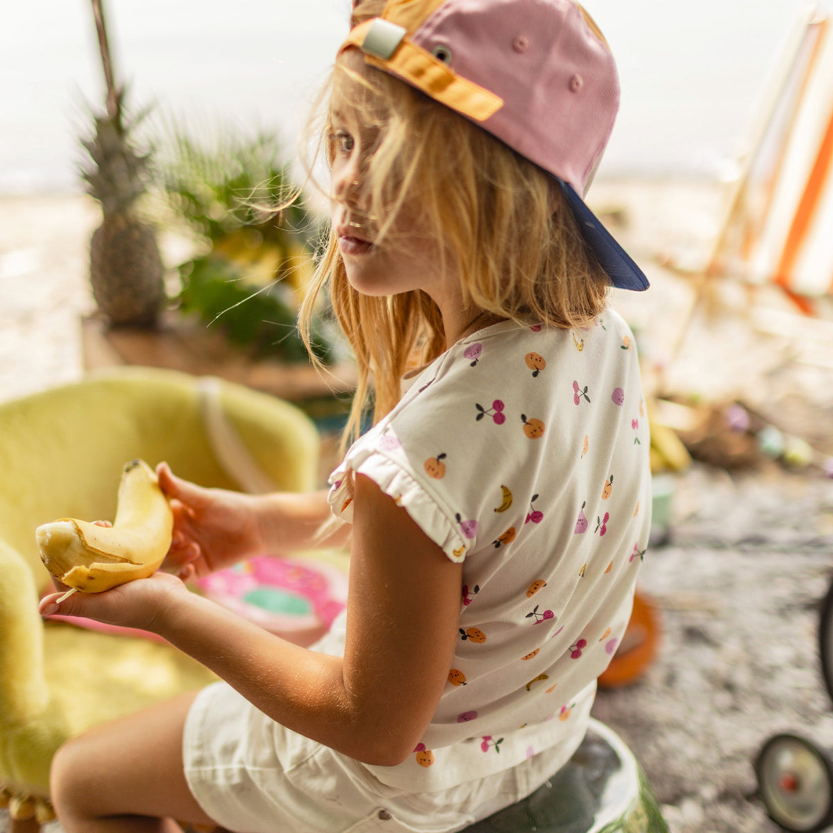 Enfant portant un haut blanc à motifs de fruits colorés, assise dehors avec une banane à la main et une casquette bicolore. - Souris Mini