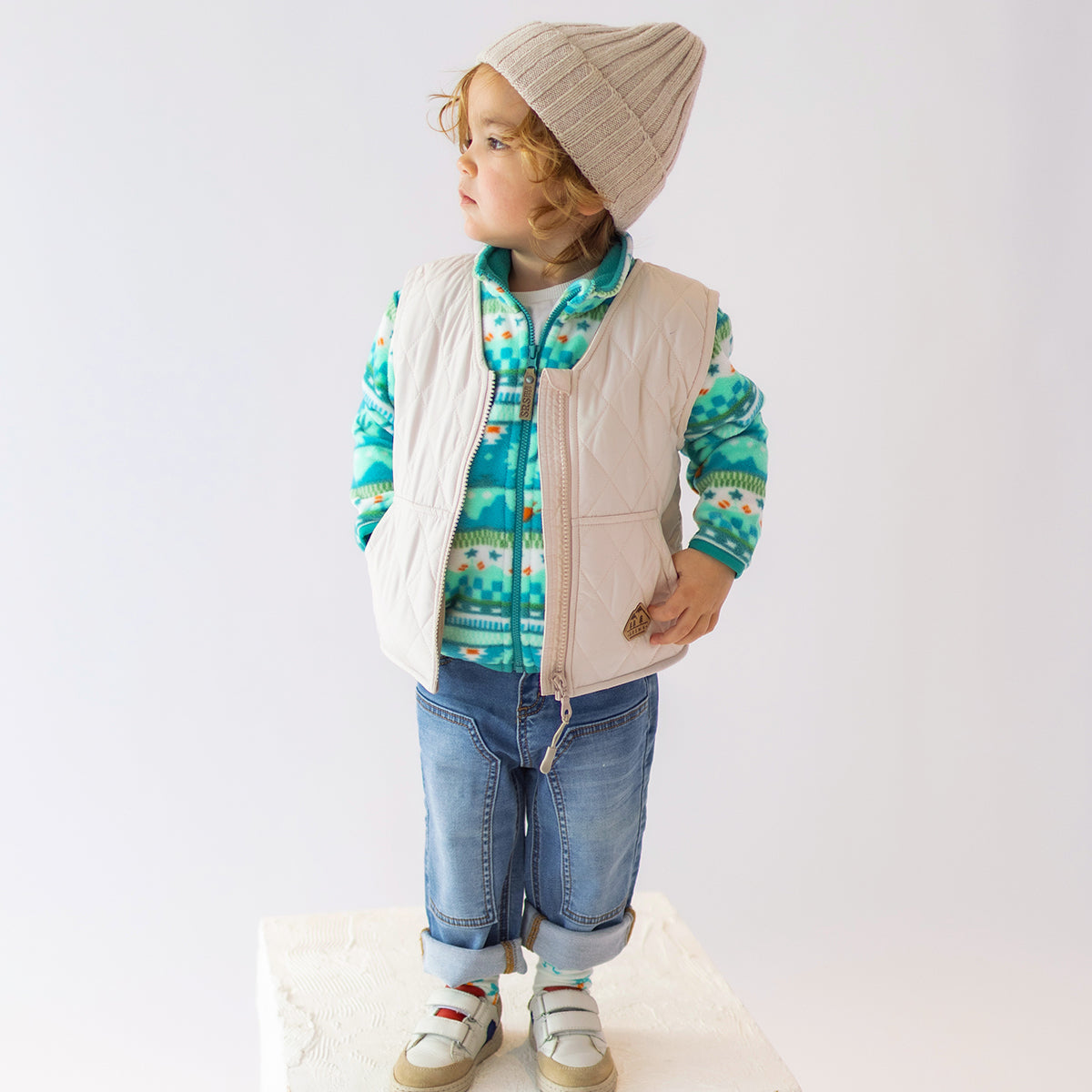 Enfant portant une polaire à motifs verts Souris Mini avec une doudoune sans manches beige et un bonnet.