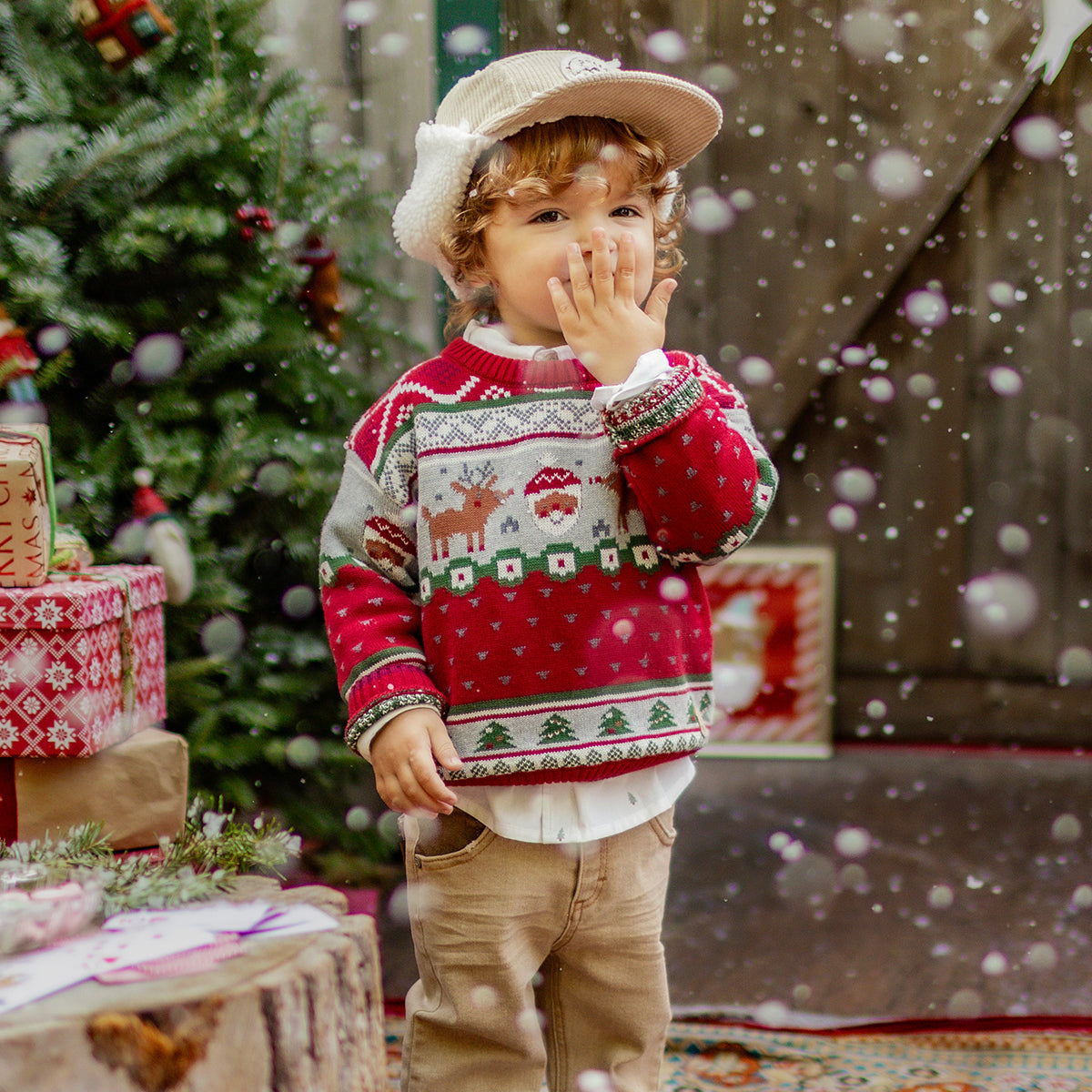 Petit garçon portant un pull rouge de Noël orné de motifs de Père Noël, de rennes et de sapins, posant dans un décor festif enneigé, collection hiver Souris Mini.