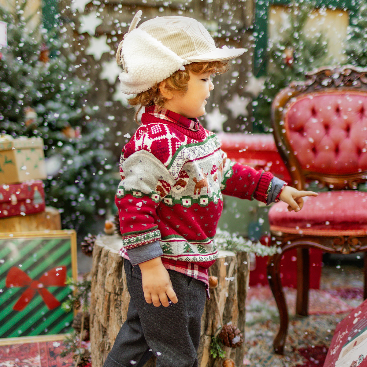 Petit garçon portant un pull tricoté rouge de Noël avec des motifs de rennes et de Père Noël, jouant sous la neige dans un décor festif avec sapin et cadeaux, collection Souris Mini.