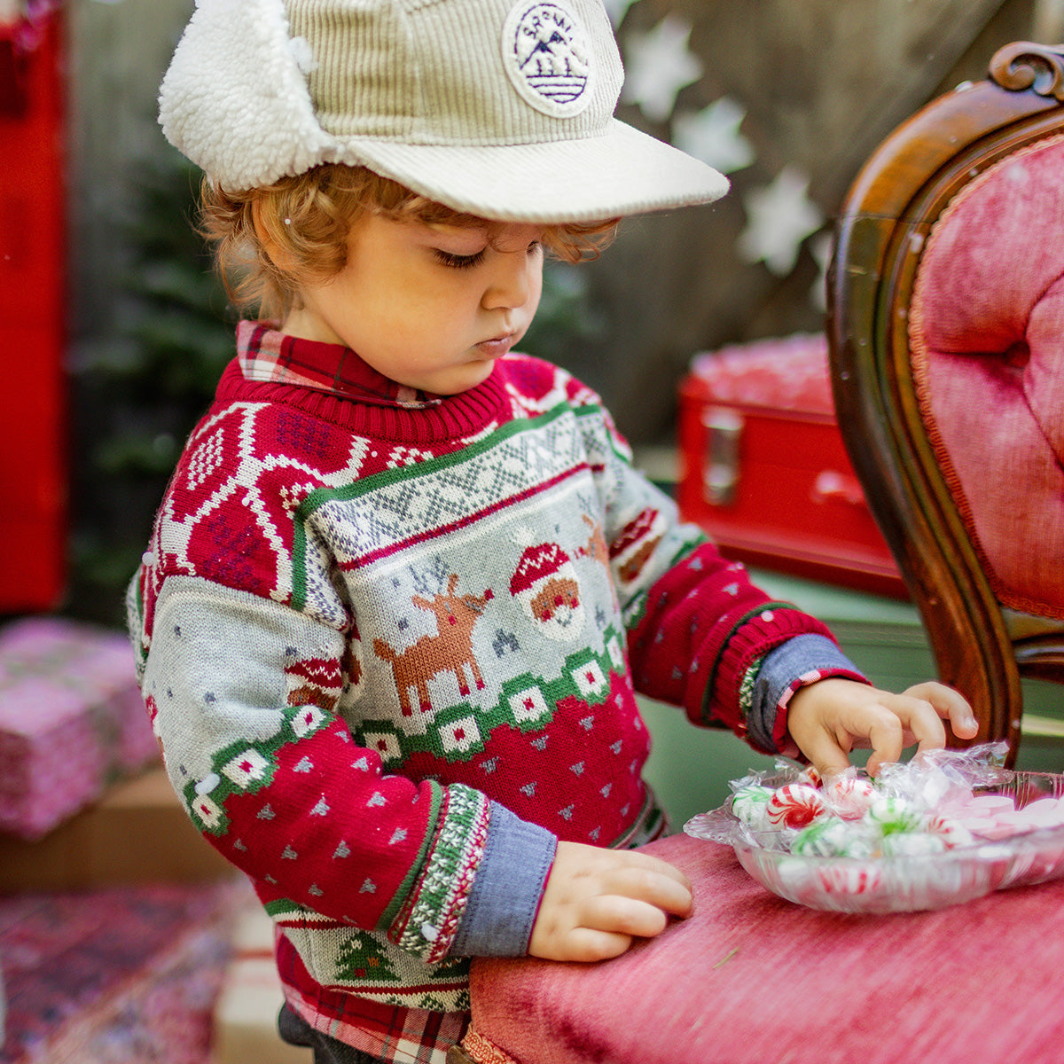 Petit garçon portant un pull tricoté rouge et gris à motifs de Noël avec rennes et Père Noël, observant des bonbons sur une chaise, ambiance festive hivernale, collection Souris Mini.