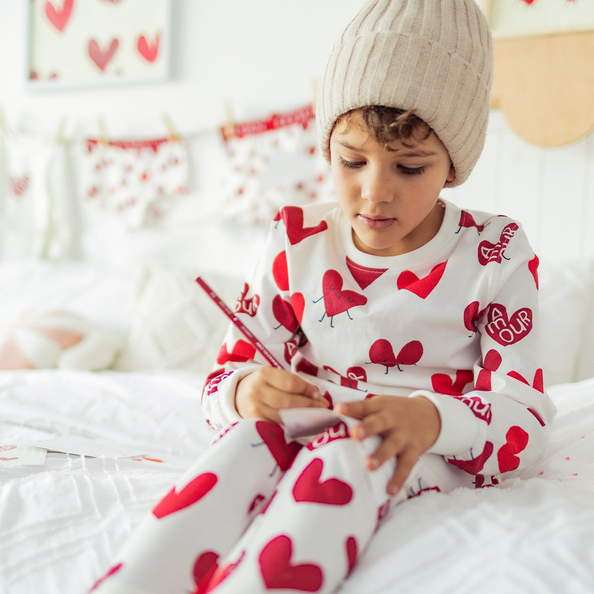 Enfant portant un pyjama blanc à motifs de cœurs rouges Souris Mini, assis sur un lit et dessinant calmement.