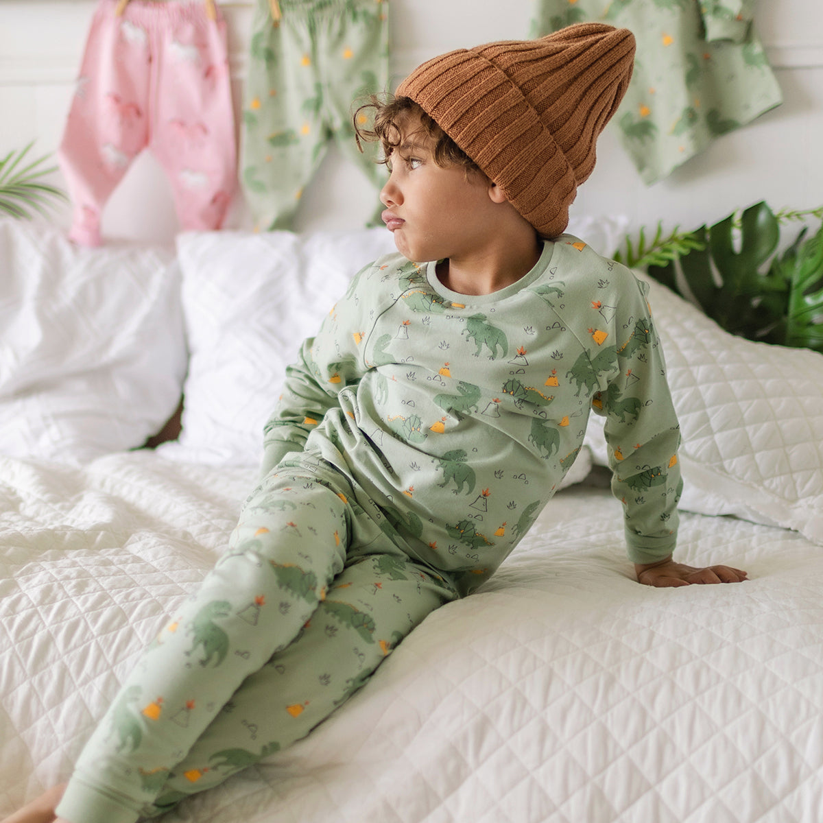 Garçon portant un pyjama vert à motifs de dinosaures Souris Mini, assis sur un lit blanc et regardant sur le côté.