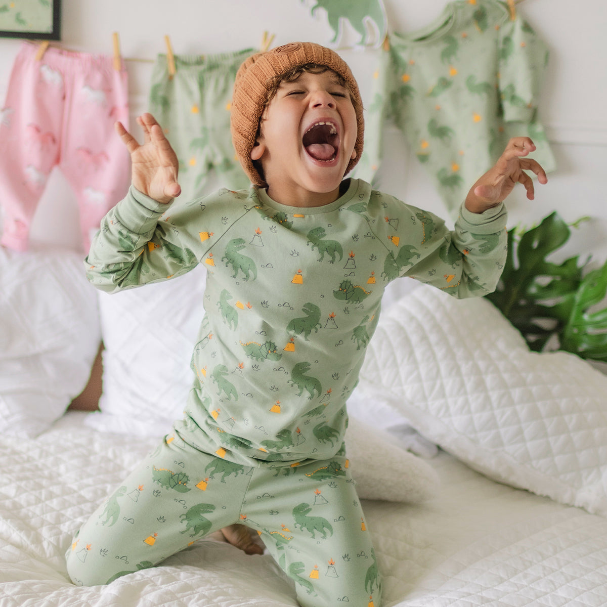 Garçon portant un pyjama vert à motifs de dinosaures Souris Mini, sautant sur un lit blanc et exprimant sa joie.