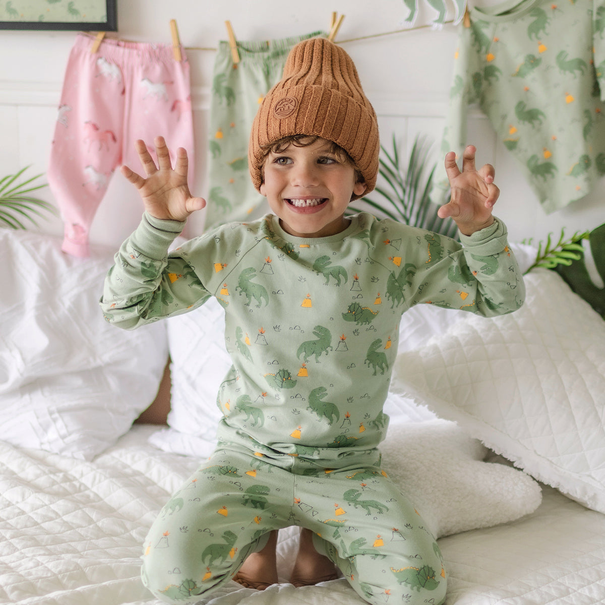 Garçon souriant portant un pyjama vert à motifs de dinosaures Souris Mini, faisant semblant de rugir sur un lit blanc.