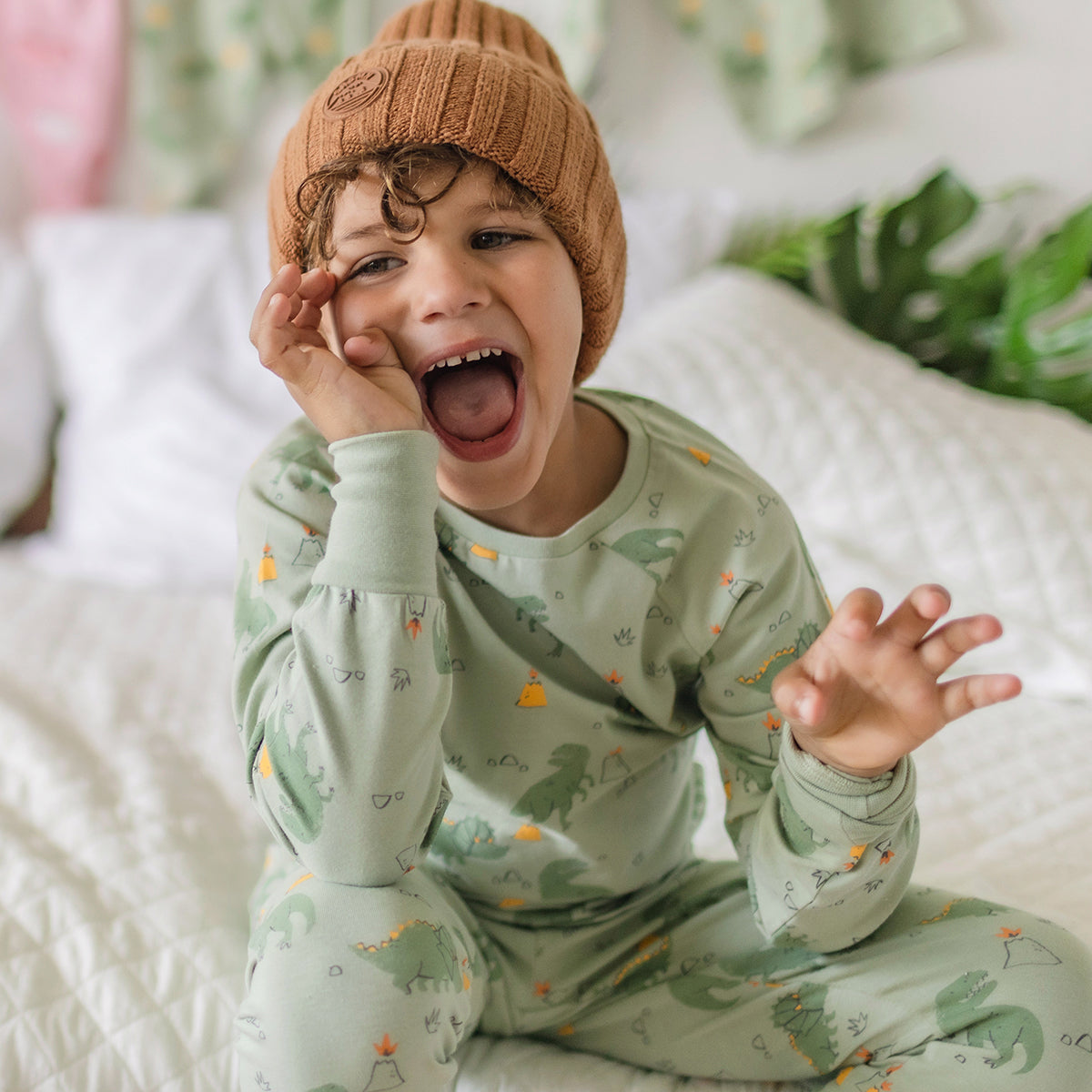 Garçon souriant portant un pyjama vert à motifs de dinosaures Souris Mini, assis sur un lit blanc avec un bonnet brun.