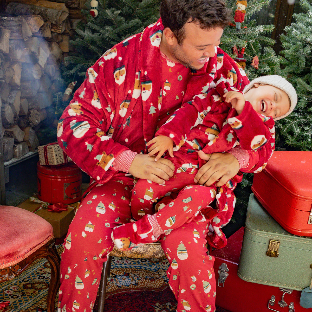 Parent et enfant en pyjamas rouges à motifs festifs rient ensemble dans un décor de Noël chaleureux avec sapins et valises rétro. - Souris Mini