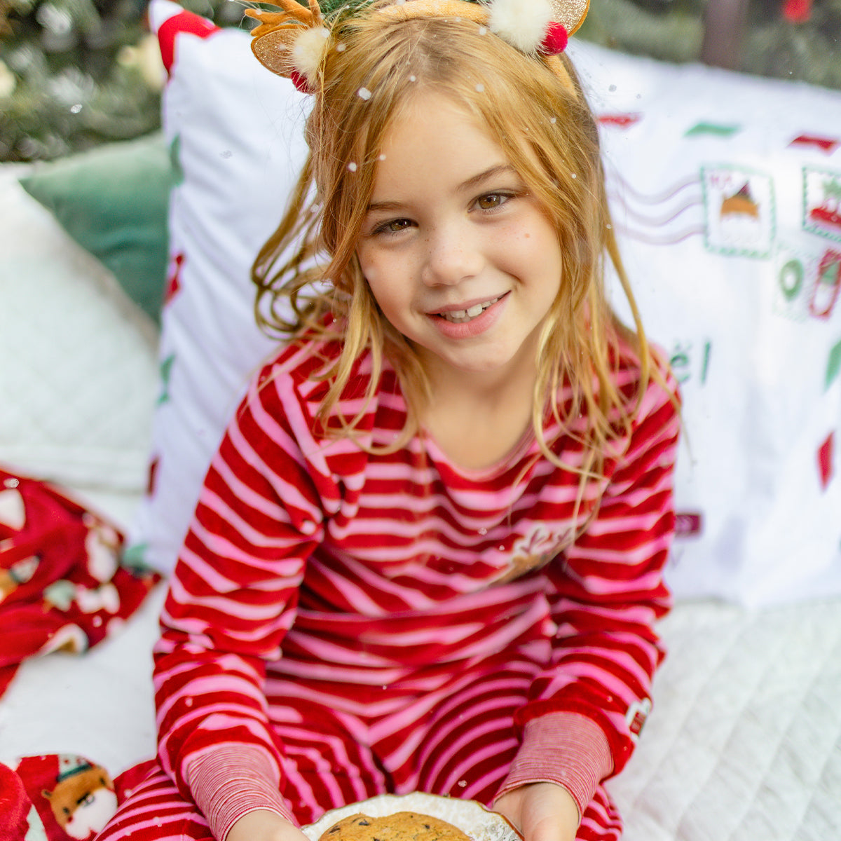 Fillette en pyjama de Noël rouge à rayures tenant une assiette de biscuits, souriant dans une ambiance festive et chaleureuse.