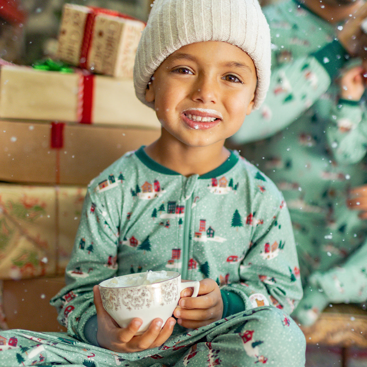 Enfant portant un pyjama vert à motifs de Noël, souriant en dégustant un chocolat chaud devant des cadeaux et un décor festif.
