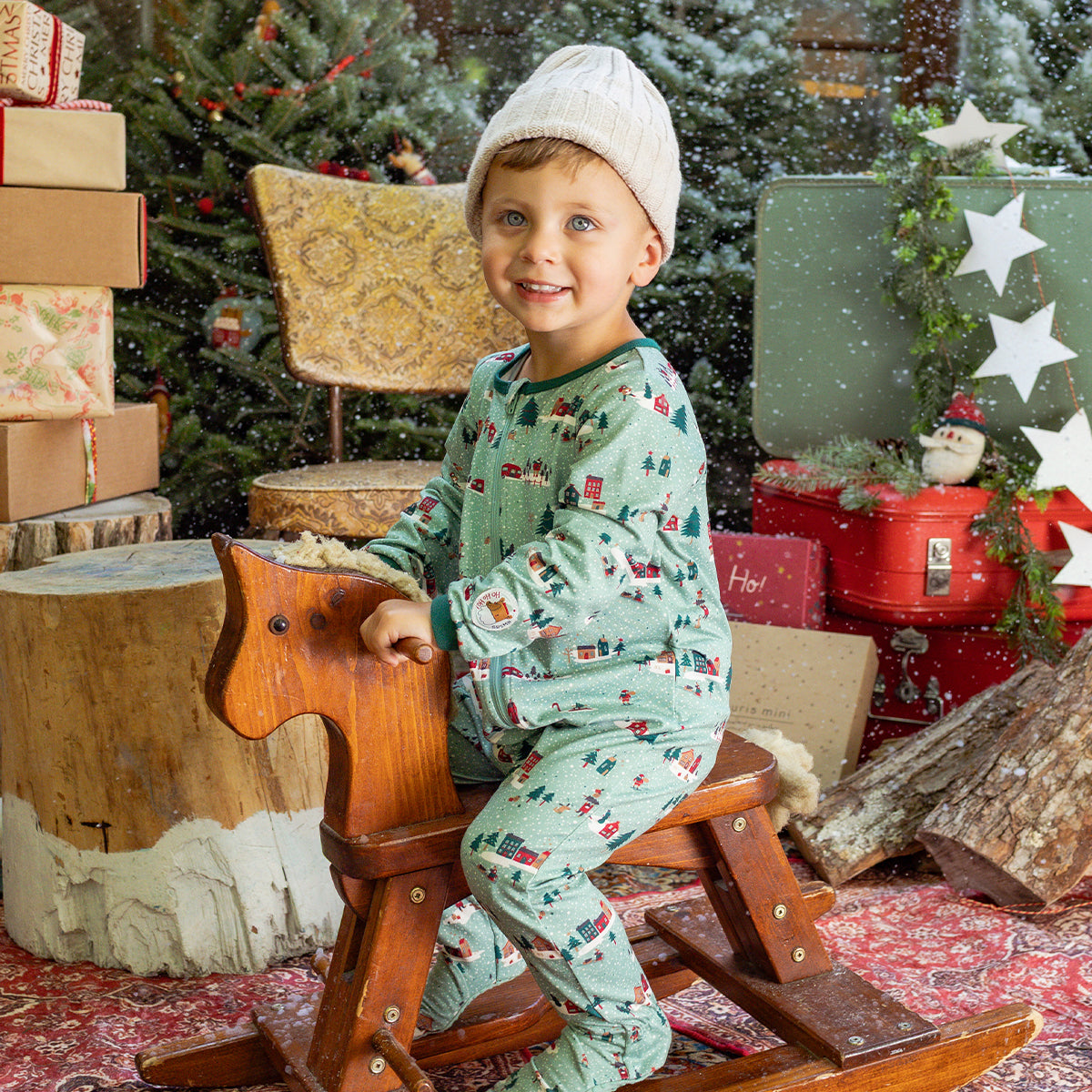 Enfant en pyjama une pièce vert à motifs de Noël assis sur un cheval à bascule, souriant dans un décor festif avec sapin et cadeaux.