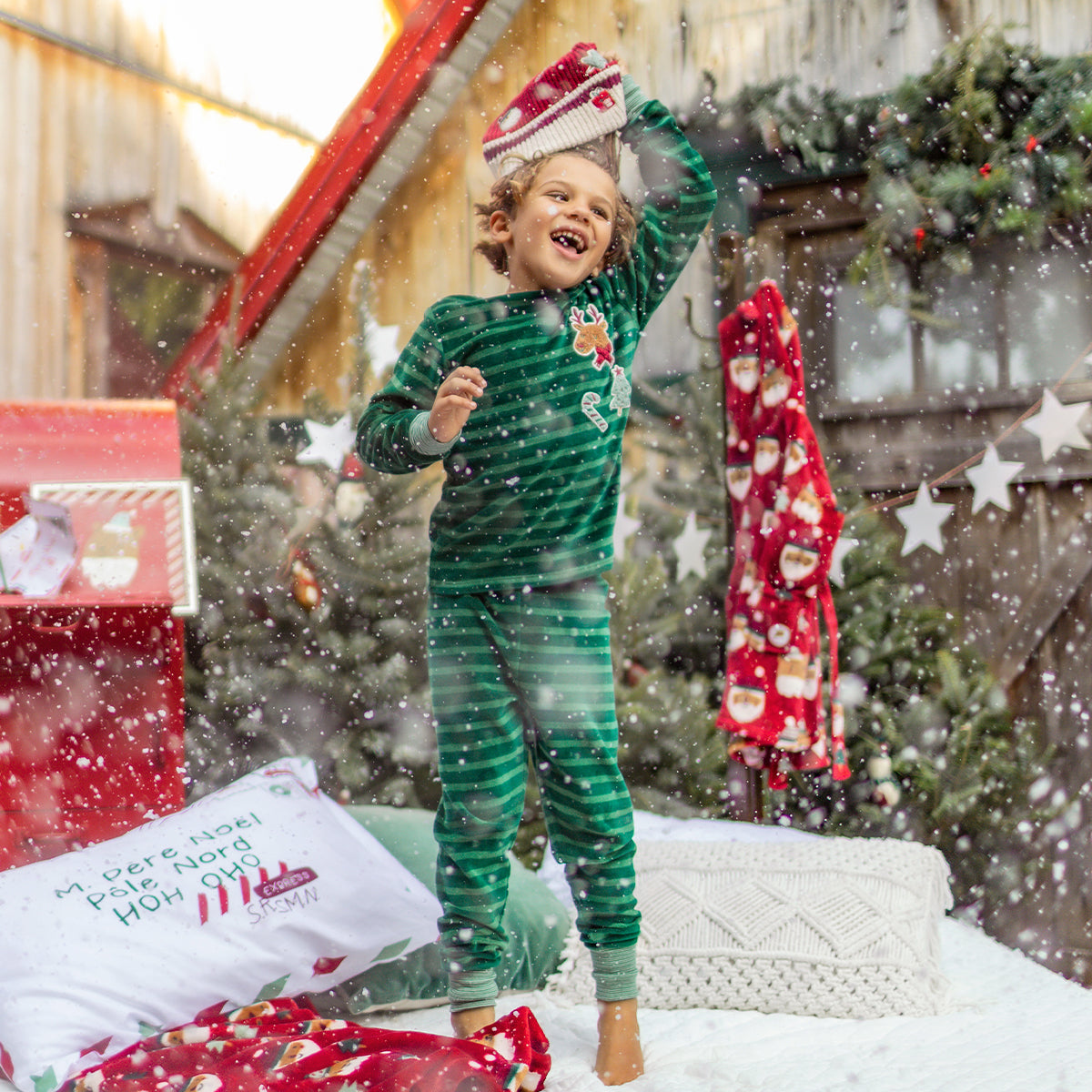 Enfant en pyjama vert à rayures et écussons festifs jouant sous la neige, riant dans un décor de Noël avec sapins et décorations rouges.