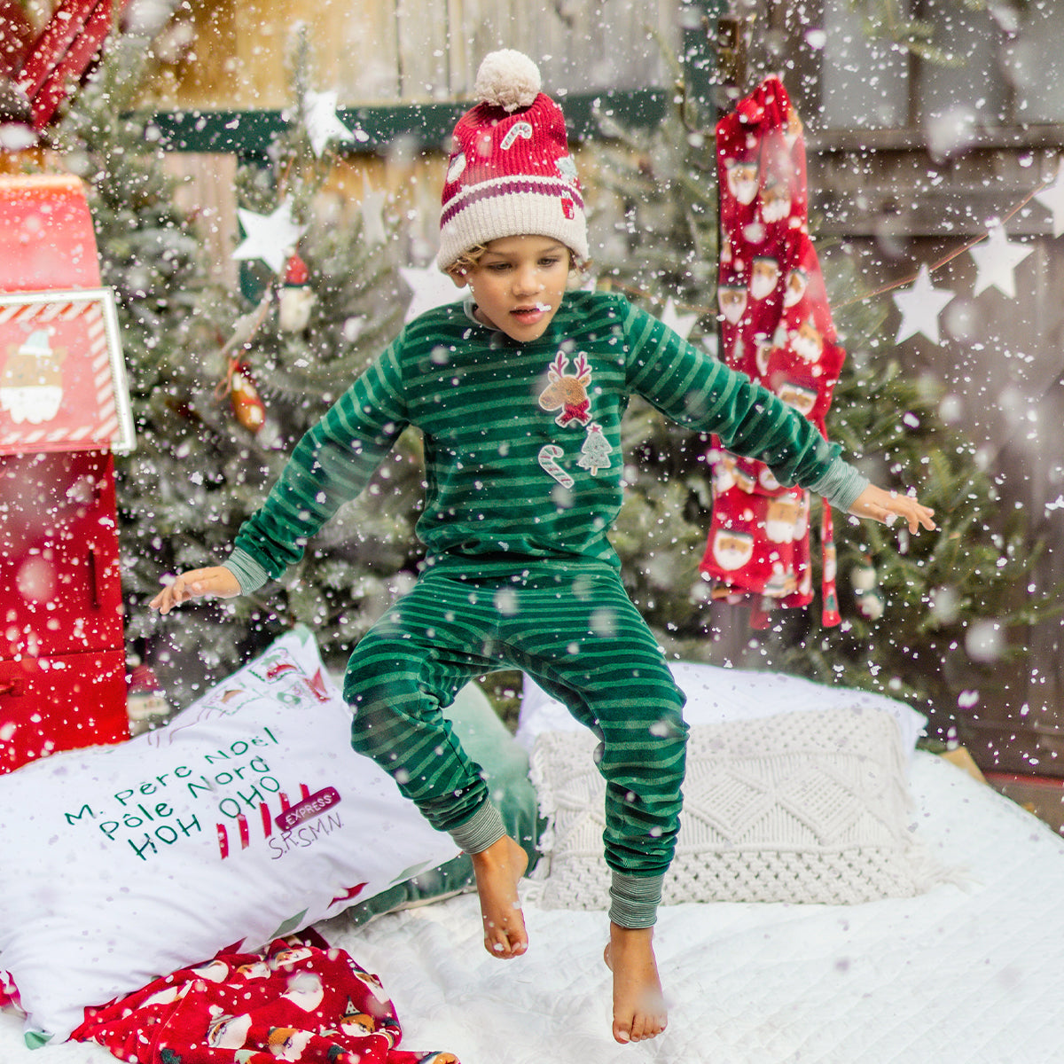 Enfant en pyjama vert à rayures et écussons festifs sautant sur un lit enneigé, entouré de décorations de Noël et d’un sapin