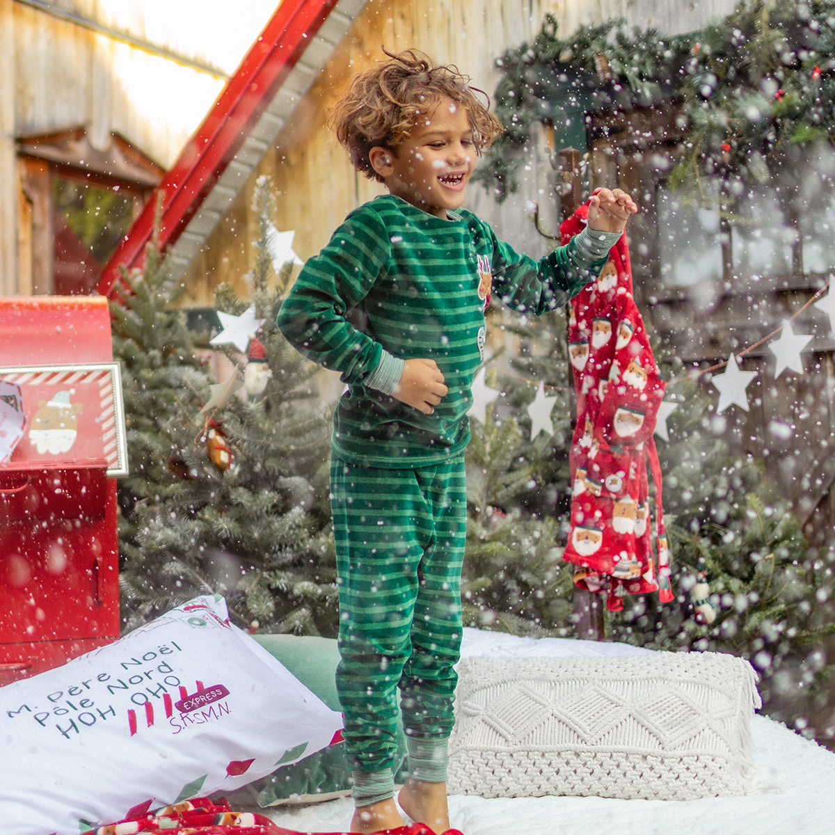 Enfant en pyjama vert à rayures et écussons festifs riant sous la neige, entouré de décorations de Noël et d’un sapin.