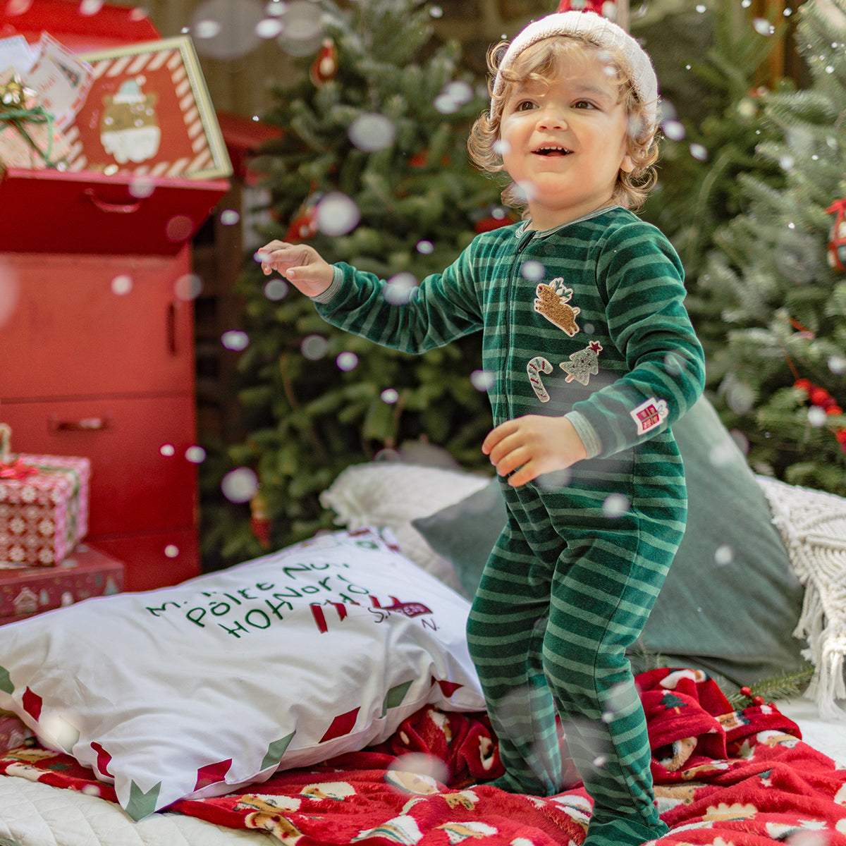 Enfant portant un pyjama une pièce vert à rayures et motifs de Noël, jouant joyeusement sous la neige dans une ambiance festive.