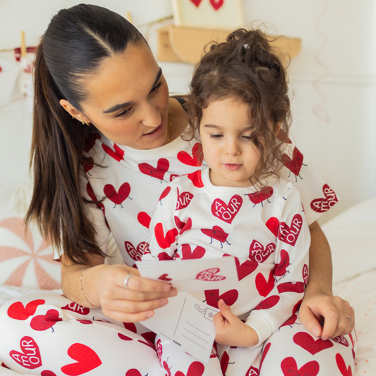 Maman et jeune enfant portant des pyjamas blancs à motifs de cœurs rouges Souris Mini, assis ensemble sur un lit et lisant une carte.