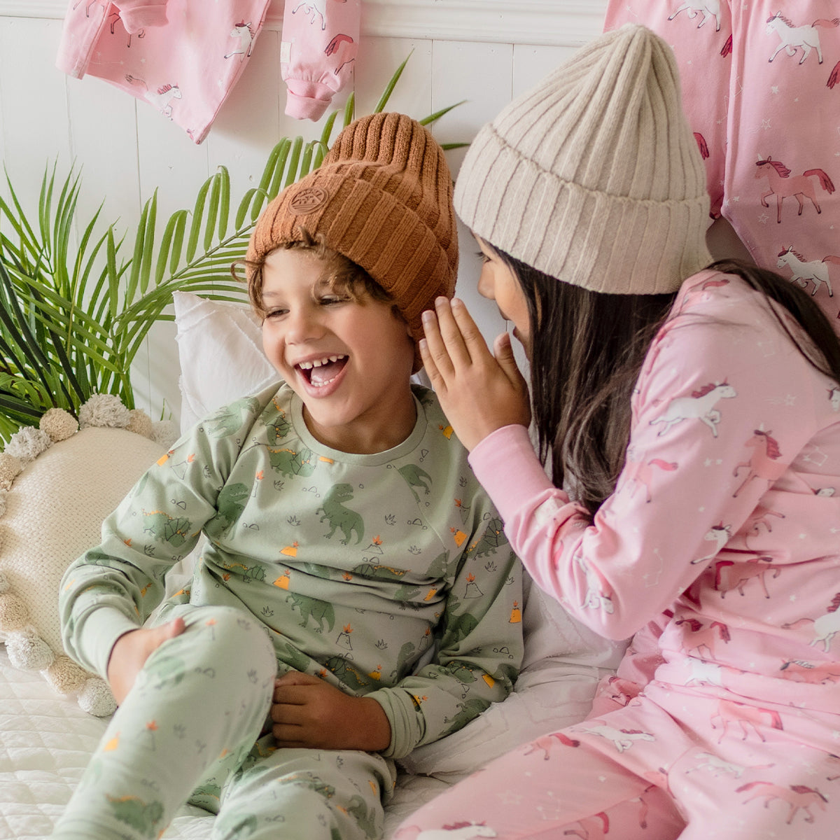 Deux enfants portant des pyjamas Souris Mini, un garçon en pyjama vert à dinosaures et une fille en pyjama rose à licornes, rient ensemble sur un lit blanc.