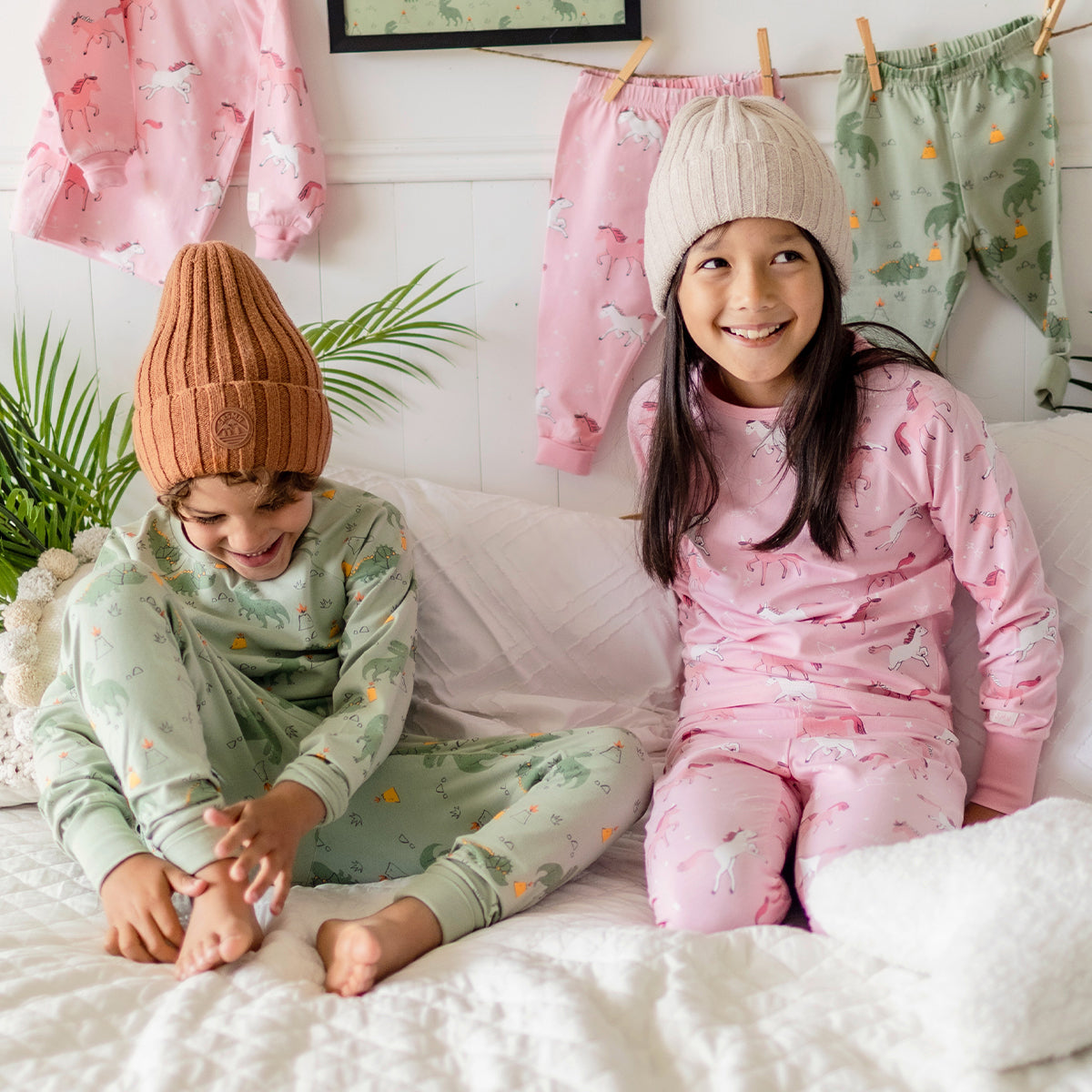 Deux enfants portant des pyjamas Souris Mini, un garçon en pyjama vert à dinosaures et une fille en pyjama rose à licornes, rient ensemble sur un lit blanc.